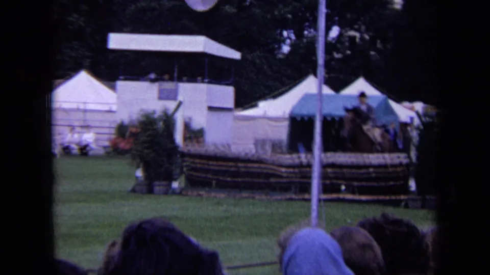 1963 London 8mm frame of an equestrian event area with a jump, white tents, spectators, and a mounted rider near frame edge.