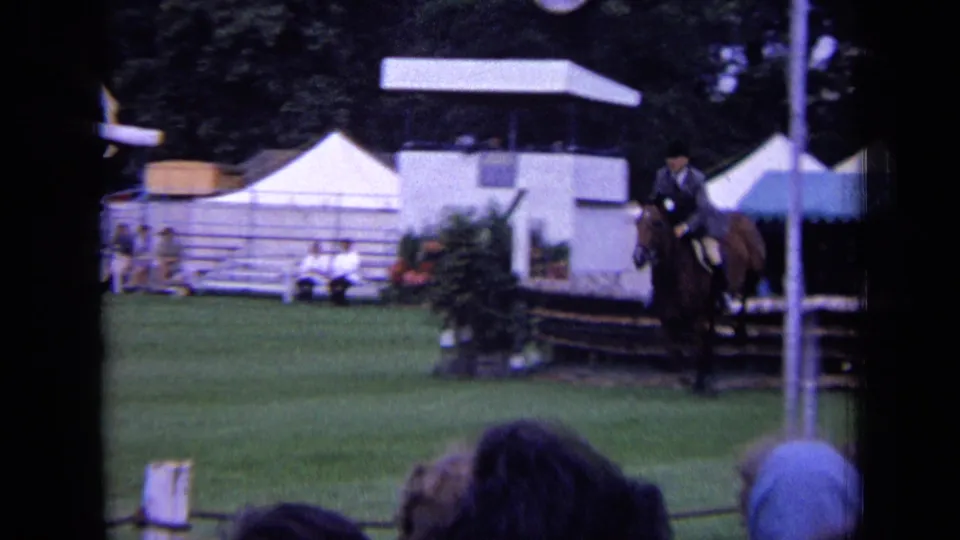 1963 London 8mm frame of a rider guiding a brown horse toward a timber jump, with tents, stand, trees, and spectators visible.