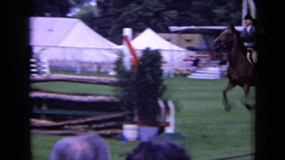 Rider and brown horse appear mid-jump at a London equestrian event, with white tents, rails, and course equipment behind them.