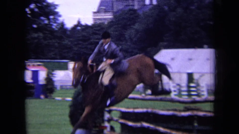 1963 London home-movie frame showing a rider on a brown horse just after a jump, with show tents and trees in the background.