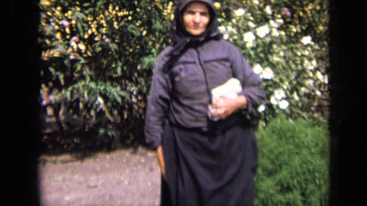 In 1963 Russia home-movie footage, an elderly woman in a dark headscarf and coat stands near garden foliage holding a white object.