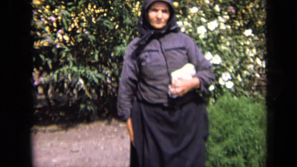 In 1963 Russia home-movie footage, an elderly woman in a dark headscarf and coat stands near garden foliage holding a white object.