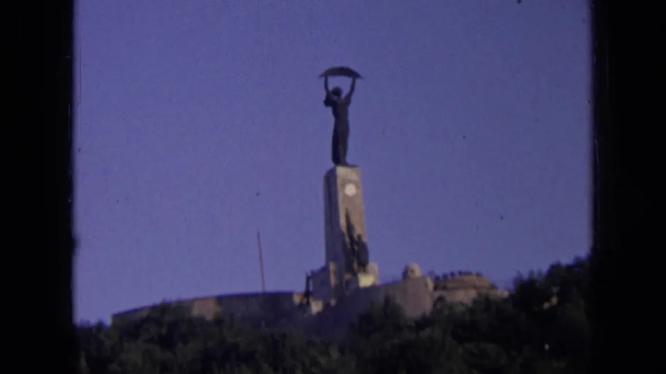 1963 home-movie frame of the distant monument in Russia, with the figure and sickle centered above trees and a low structure.