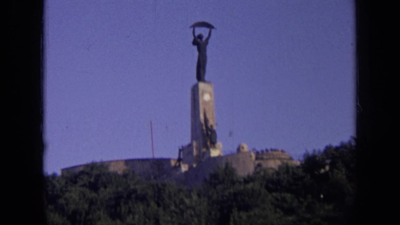 Distant statue on a tall pedestal above a wooded hillside under blue sky.