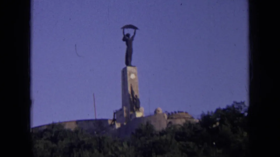 1963 home-movie frame from Russia showing the monumental statue and pedestal above dense trees, with dark film borders visible.
