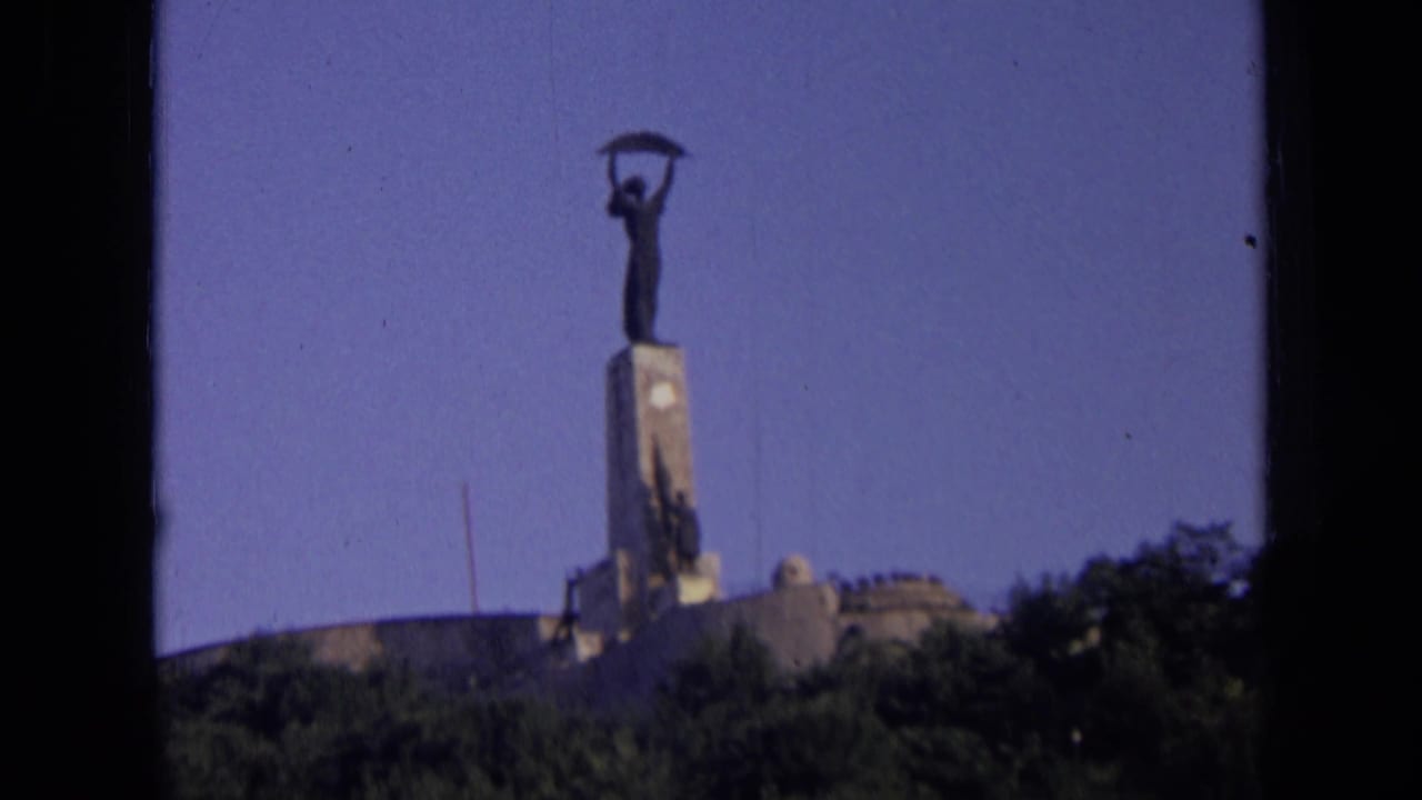 1963 Russia 8mm frame showing a distant hilltop monument with a figure holding an object aloft above trees and low buildings.