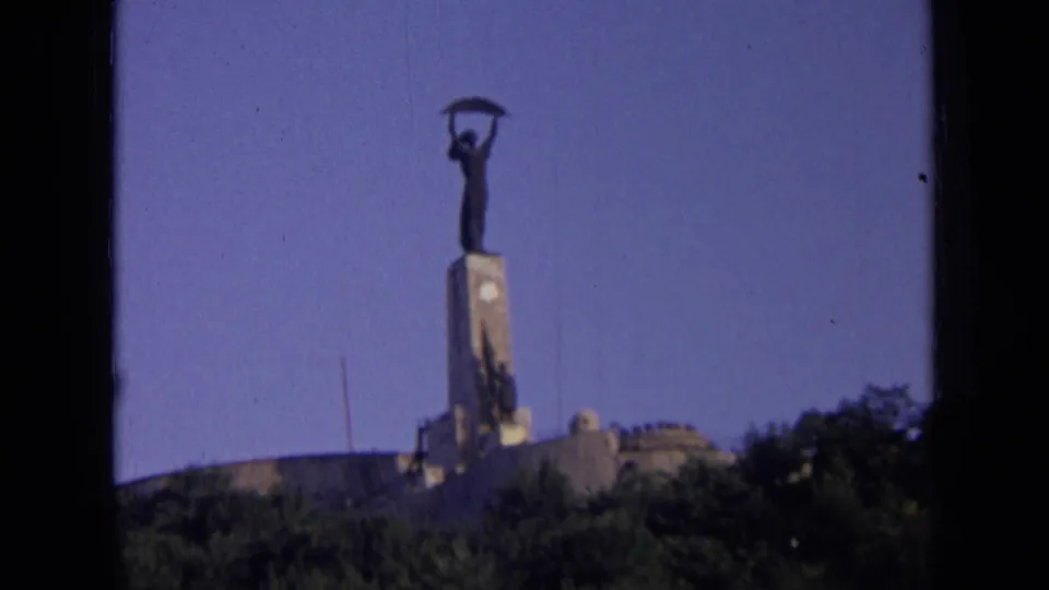 1963 Russia 8mm frame showing a distant hilltop monument with a figure holding an object aloft above trees and low buildings.