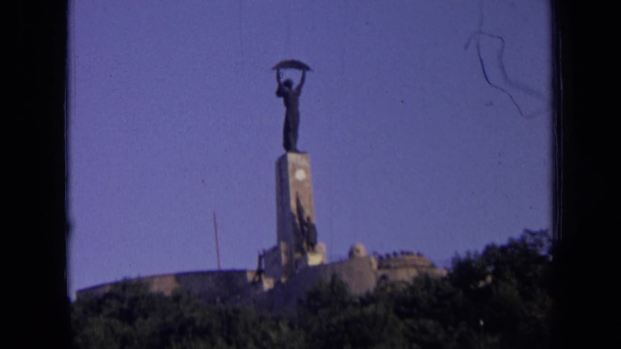 Distant 1963 Russia home-movie view of a statue on a pedestal, framed by sky, trees, and dark film gate edges.