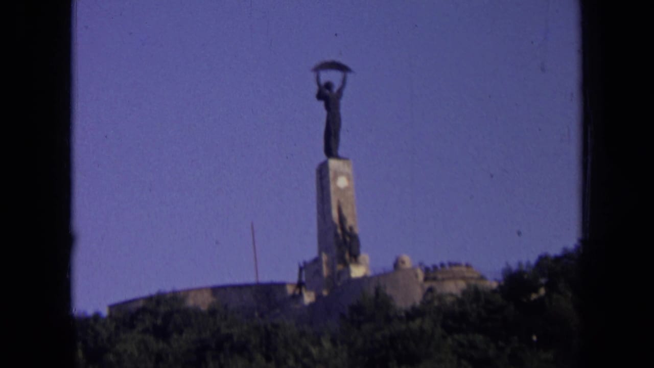 Low-angle view of a tall statue on a stone monument above trees under a blue sky.