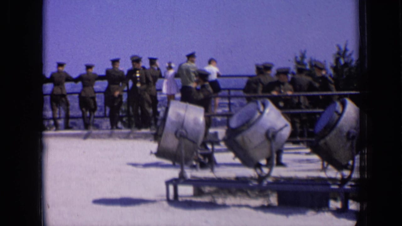 Uniformed men stand by a railing behind large tilted searchlights on an overlook.