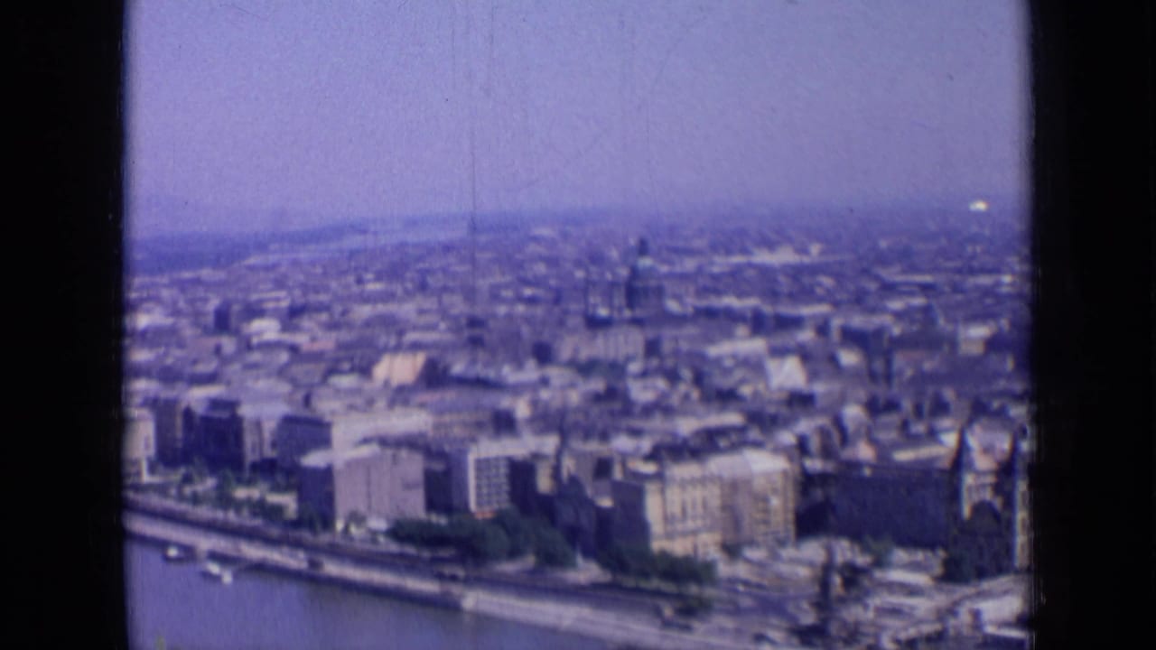 1963 8mm frame showing an elevated Russian city view across a river, with dense blocks, haze, and a distant tower-like structure.