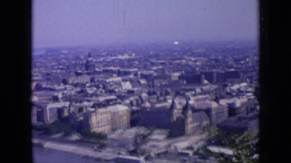 1963 8mm view over a Russian city, framed by dark film divisions, with riverbank blocks and a hazy urban horizon.
