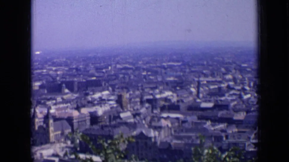 1963 8mm frame looking over a Russian city, with foreground foliage, packed rooftops, and a hazy distant skyline.