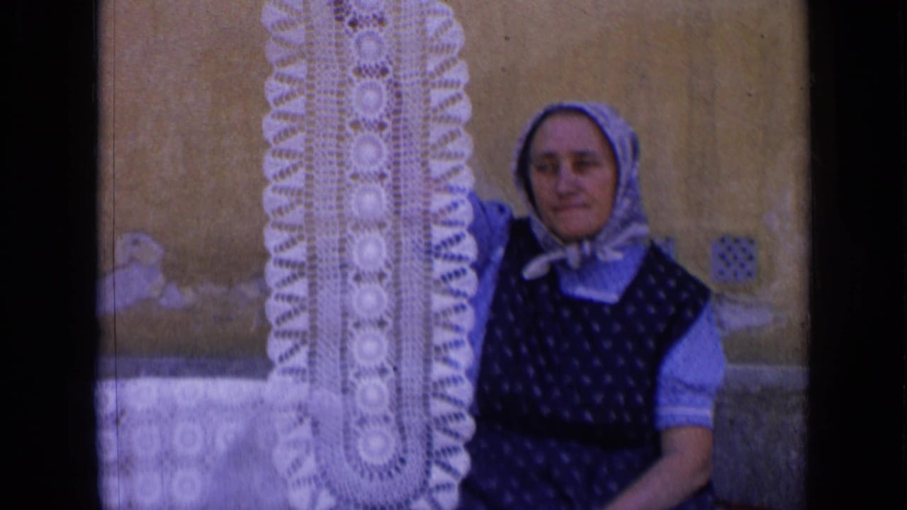 Woman in a headscarf holds a long white crocheted doily beside a worn wall.