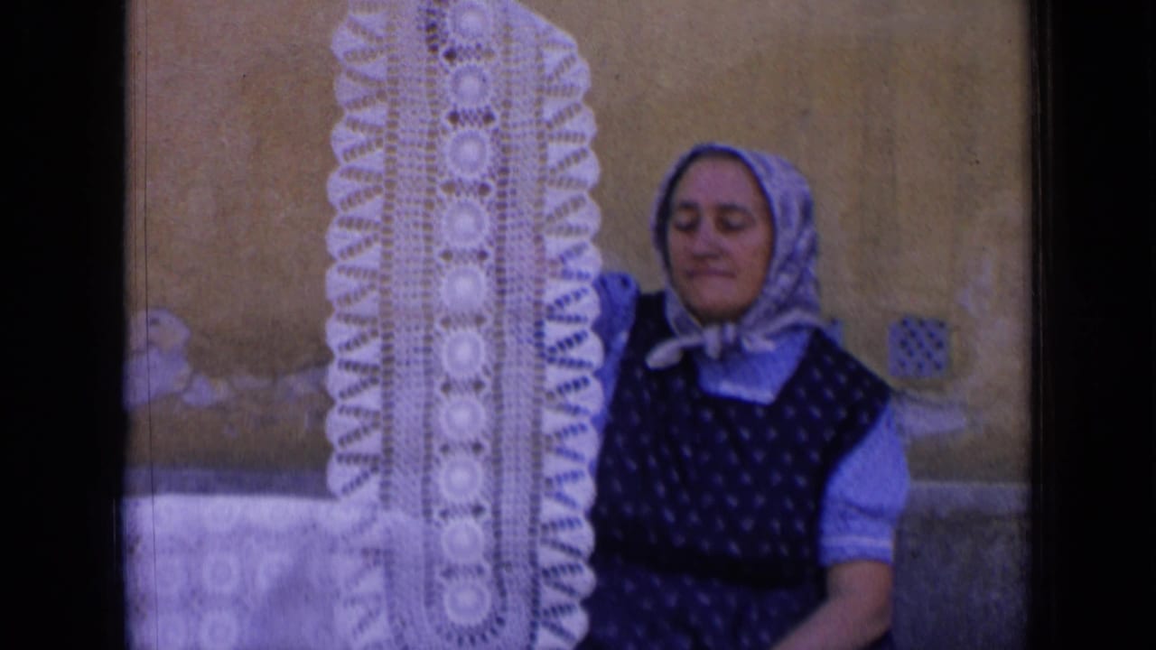 A woman in 1963 Russia footage lowers her gaze while holding a tall white crocheted doily beside a wall.