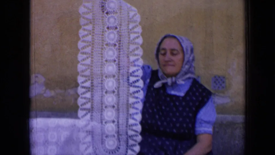 A woman in 1963 Russia footage lowers her gaze while holding a tall white crocheted doily beside a wall.