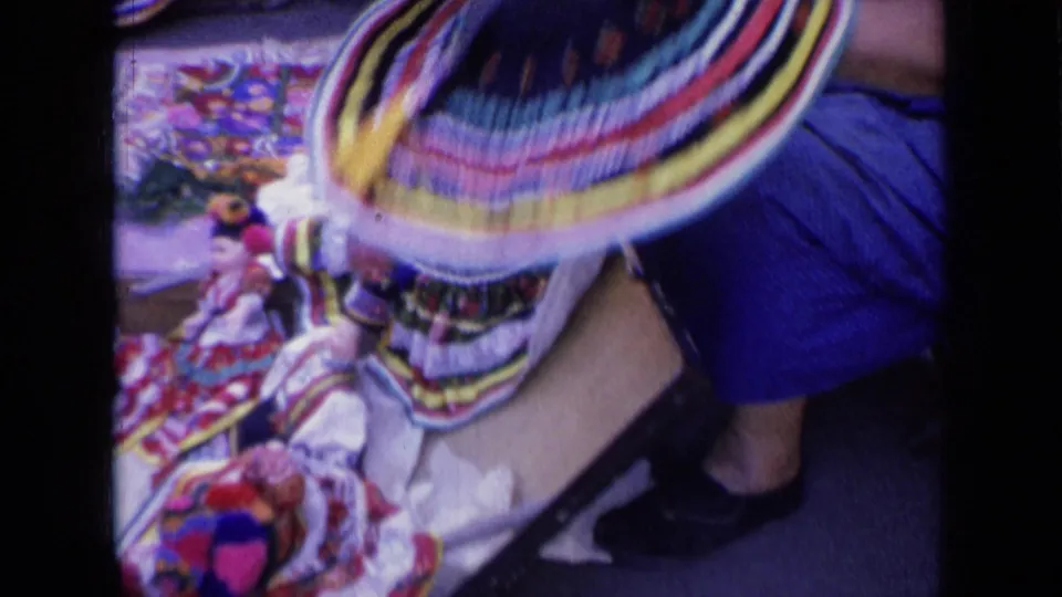 The clip shifts to colorful crocheted or lace textiles, with a partial figure in blue clothing visible at the edge of the frame.