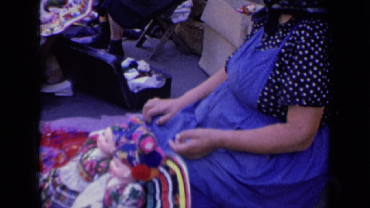 Seated woman in a blue dress beside colorful dolls.