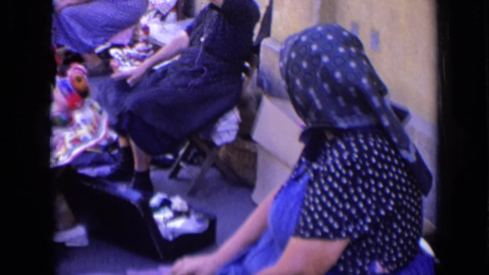 A woman wearing a headscarf sits near dolls, fabric, and a small open box in the outdoor 8mm scene.