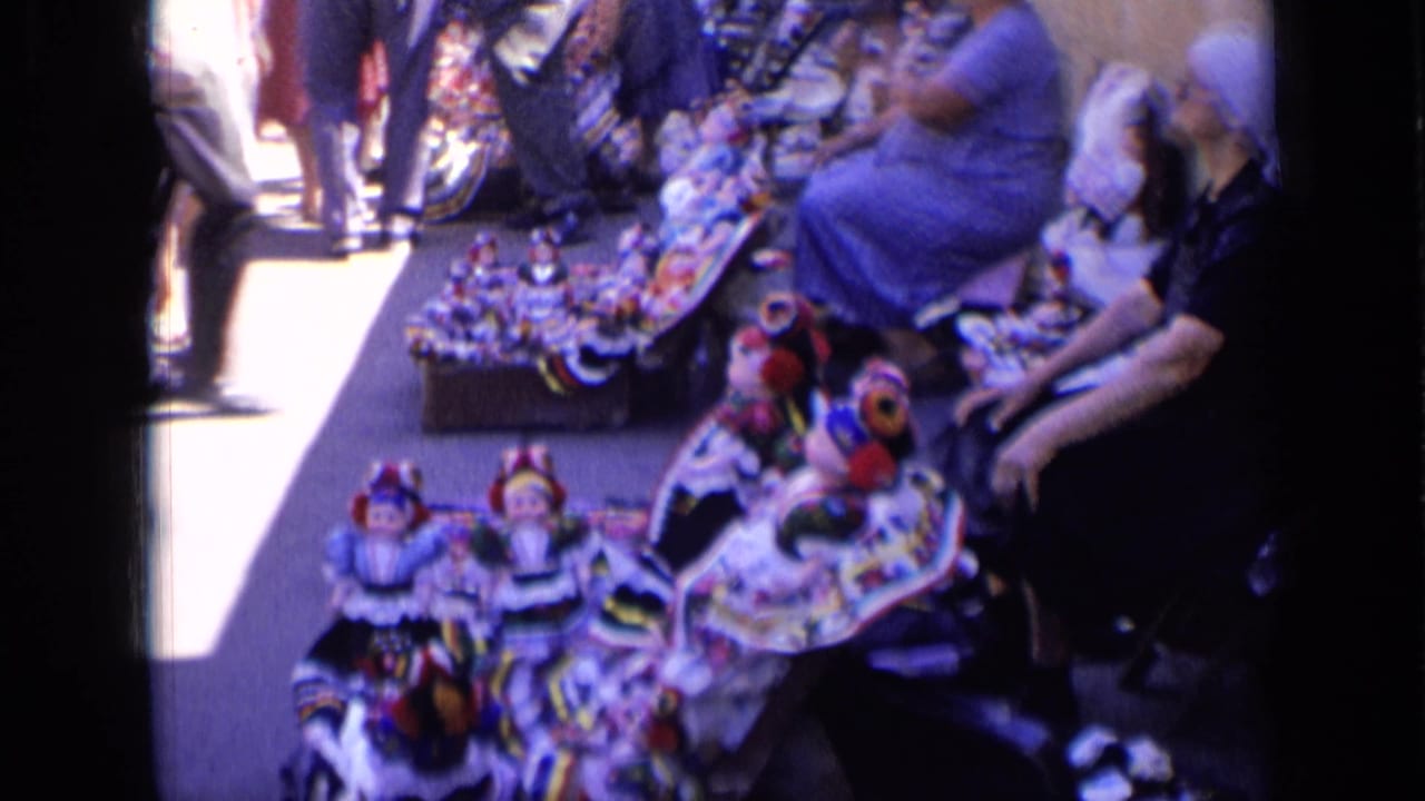 1963 Russia 8mm frame showing a woman vendor seated near rows of traditional-looking dolls on a sunlit sidewalk.
