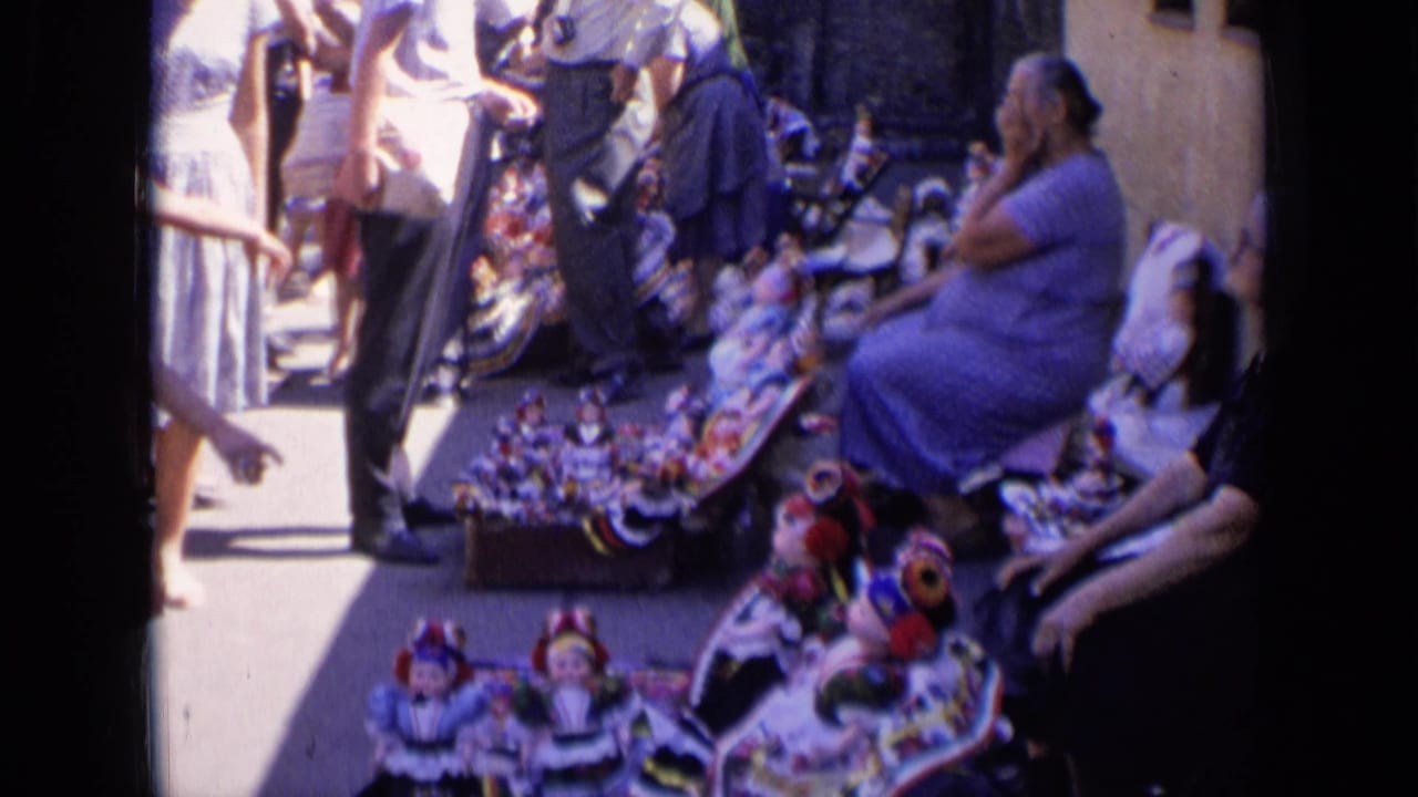 Woman seated beside dolls and craft items with passersby walking along the street.