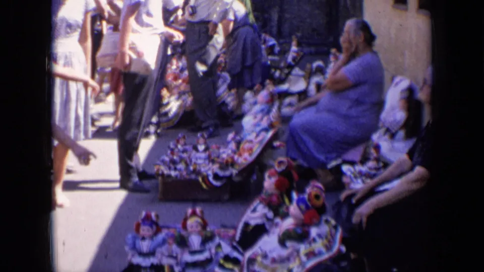 Home-movie view in Russia, 1963, with a seated woman and a bright sidewalk display of handmade folk-style dolls.