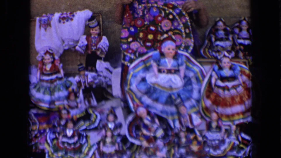 1963 Russia home-movie frame showing a blue-skirted doll at center with surrounding folk-costume figures in a dense arrangement.