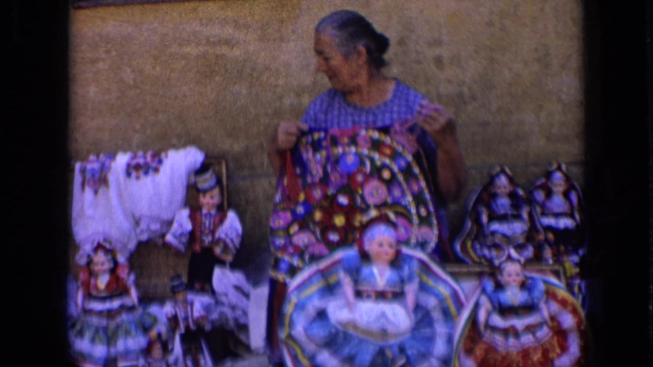 The woman stands behind a table of handmade dolls, holding a bright patterned cloth and looking across the display.