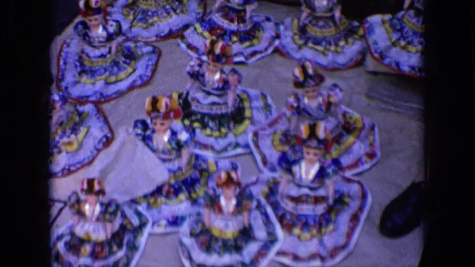 Rows of small folk-style dolls with embroidered skirts and hats fill the frame, with part of a shoe visible at the table edge.