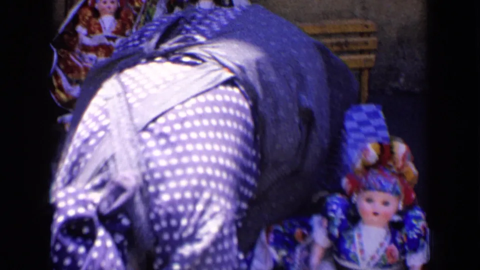 The vendor's polka-dot dress and dark scarf fill the foreground beside a handmade doll on the display.