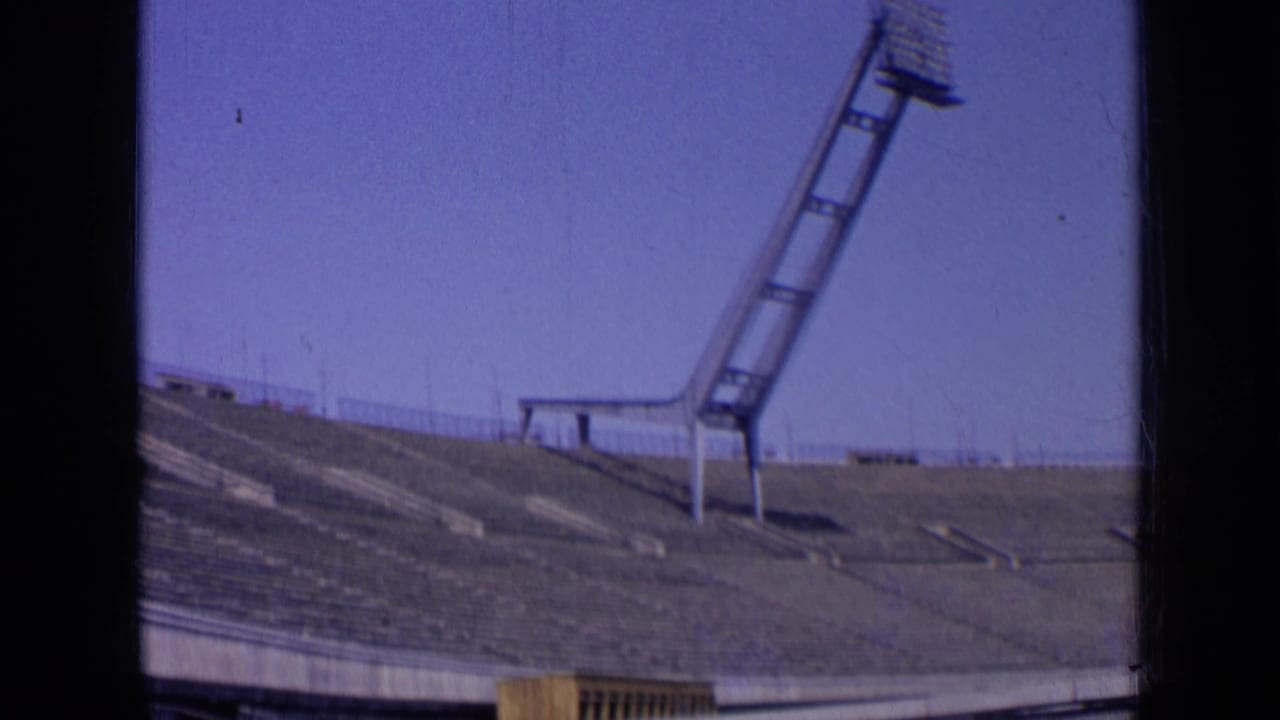 Empty stadium bleachers below a tall floodlight mast against blue sky.