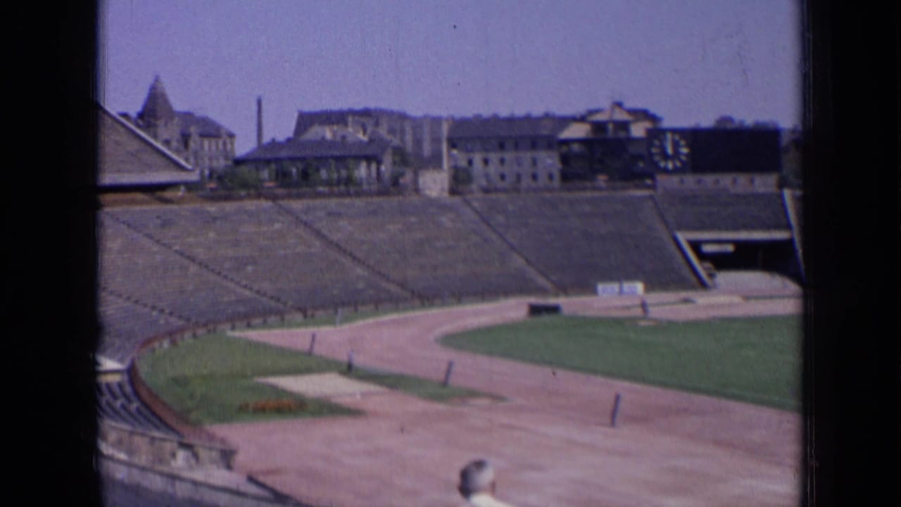 Empty stadium seating, running track, green field, and distant buildings under a bright sky.
