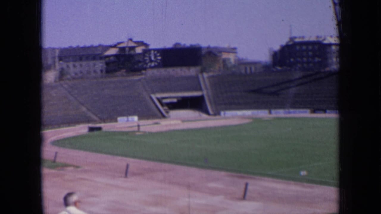 Panned 8mm frame of a Russian stadium in 1963, showing empty terraces, field, track, and distant rooflines.