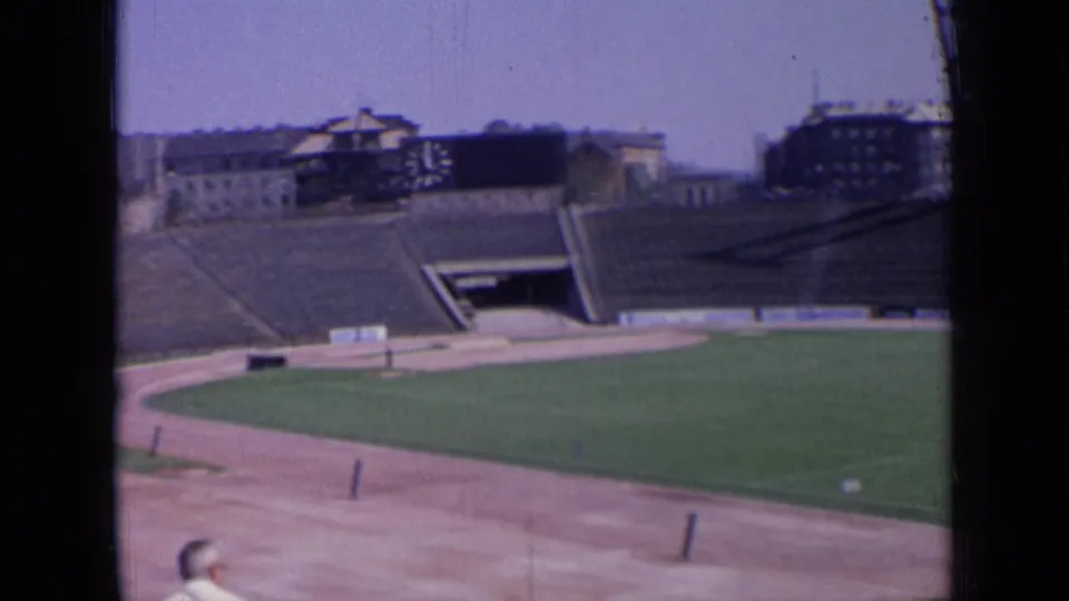 Panned 8mm frame of a Russian stadium in 1963, showing empty terraces, field, track, and distant rooflines.