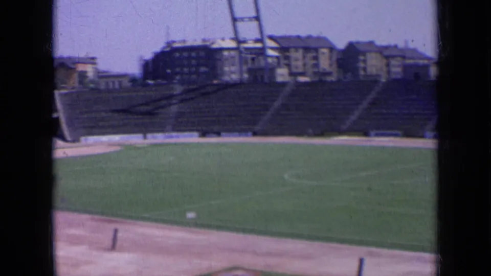 1963 Russia home-movie frame with a clock tower rising behind empty stadium seating and a green playing field.