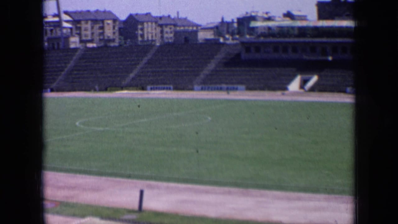 Empty grass football field with concrete stadium seating and buildings beyond.