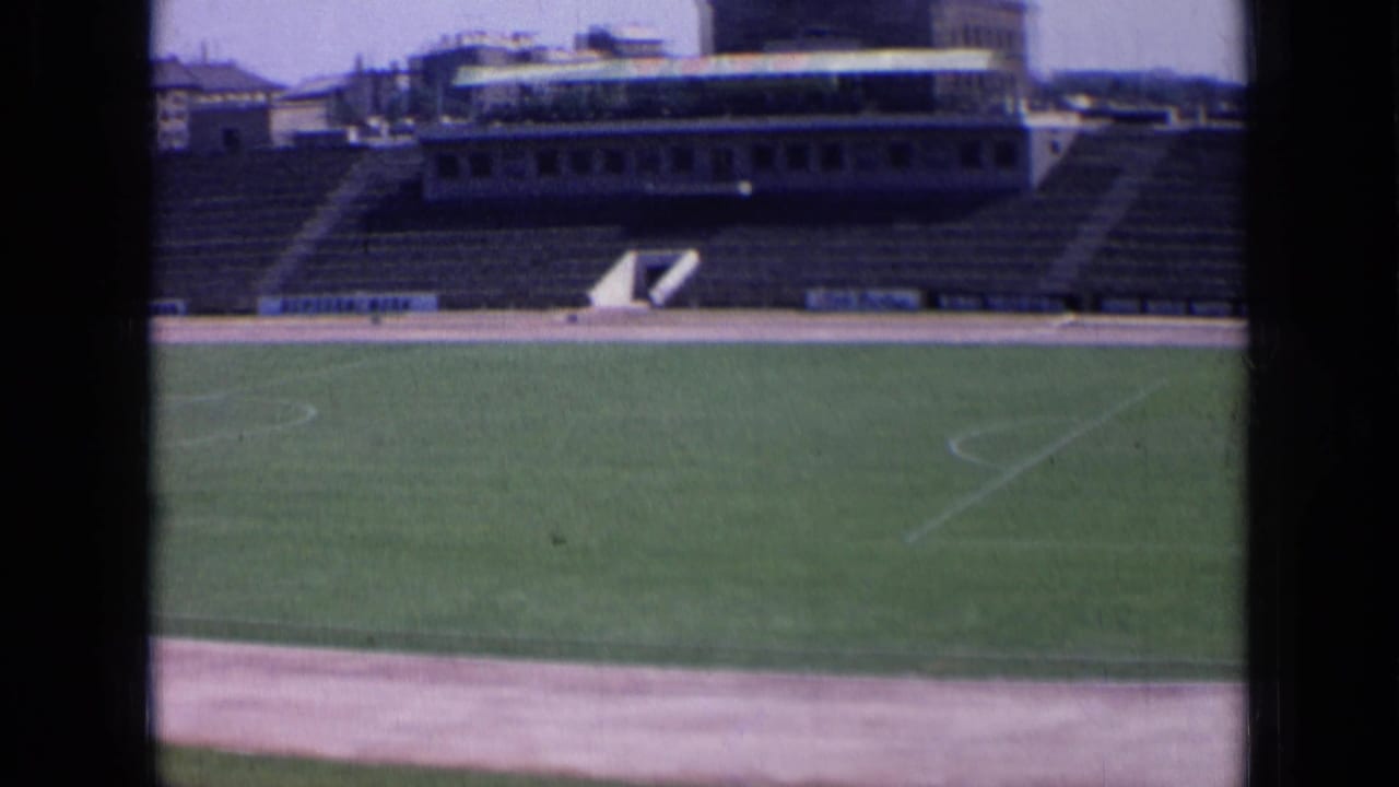 Empty Russian stadium field in 1963 footage, with white pitch lines, concrete terraces, and surrounding buildings.