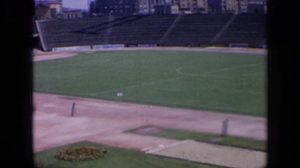 1963 Russia 8mm frame showing a stadium field and track, with a small flower bed, empty seating, and buildings beyond.