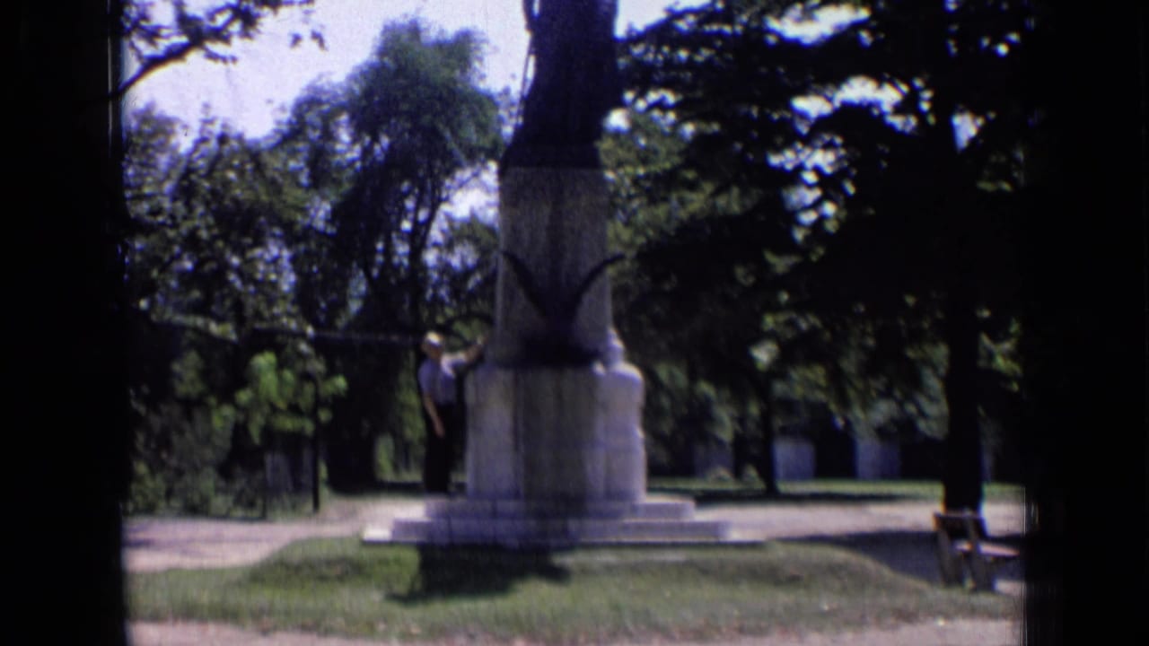 Person standing beside a stone monument in a sunny park with trees and grass.
