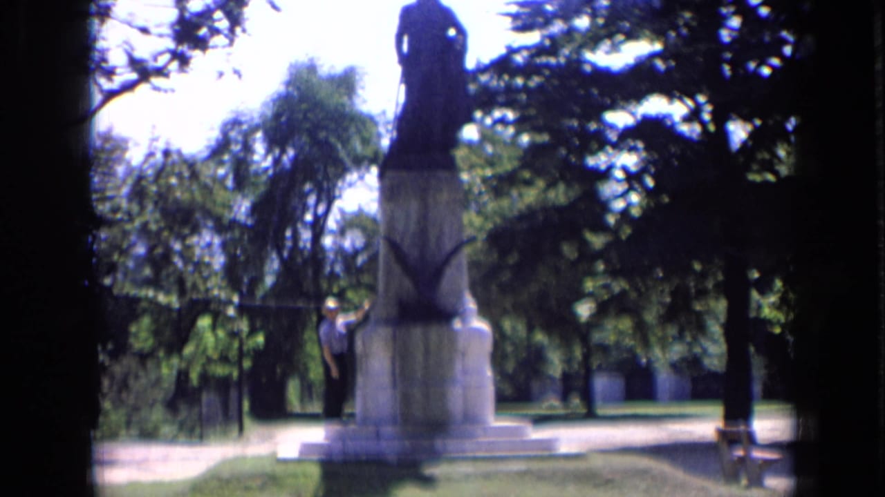 1963 Russia frame with the Dzerzhinsky monument centered, a visitor at the base, trees, grass, and bright daylight around the park path.