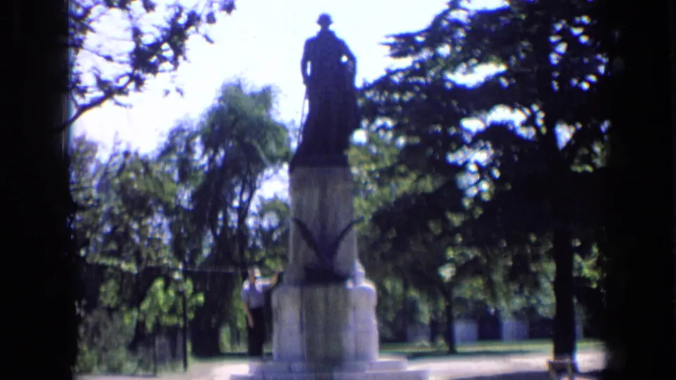 1963 Russia home-movie view of the Dzerzhinsky monument, with a visitor beside the pedestal and dark foliage framing the sunny park scene.