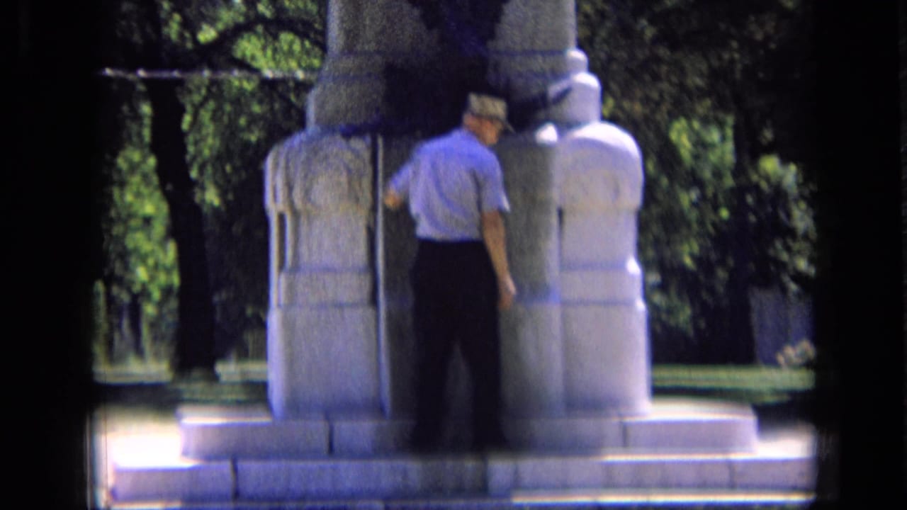 Man in short sleeves and a cap studies the base of a stone memorial in 1963 Russia home-movie footage.