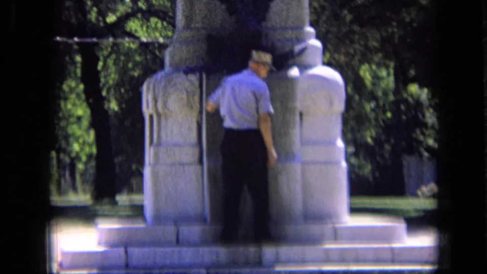 Man in short sleeves and a cap studies the base of a stone memorial in 1963 Russia home-movie footage.