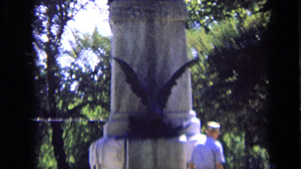 Close 8mm view of the memorial base with a dark sculpted bird-like shape and a visitor at the side.