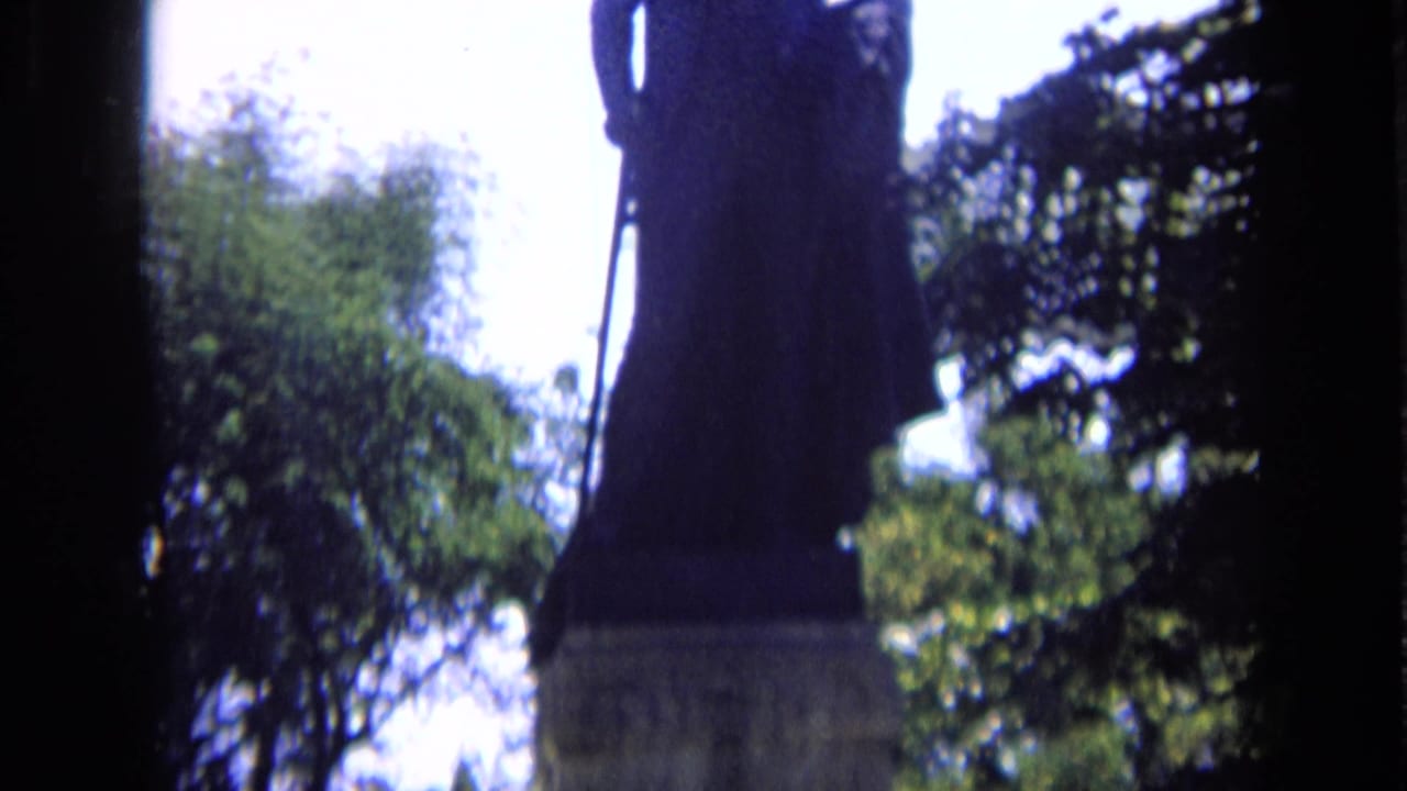 Backlit statue partly cropped among trees with bright sky behind it.