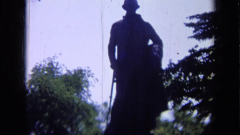 1963 Russia 8mm frame of a backlit standing statue in a hat, with trees and pale sky in the background.