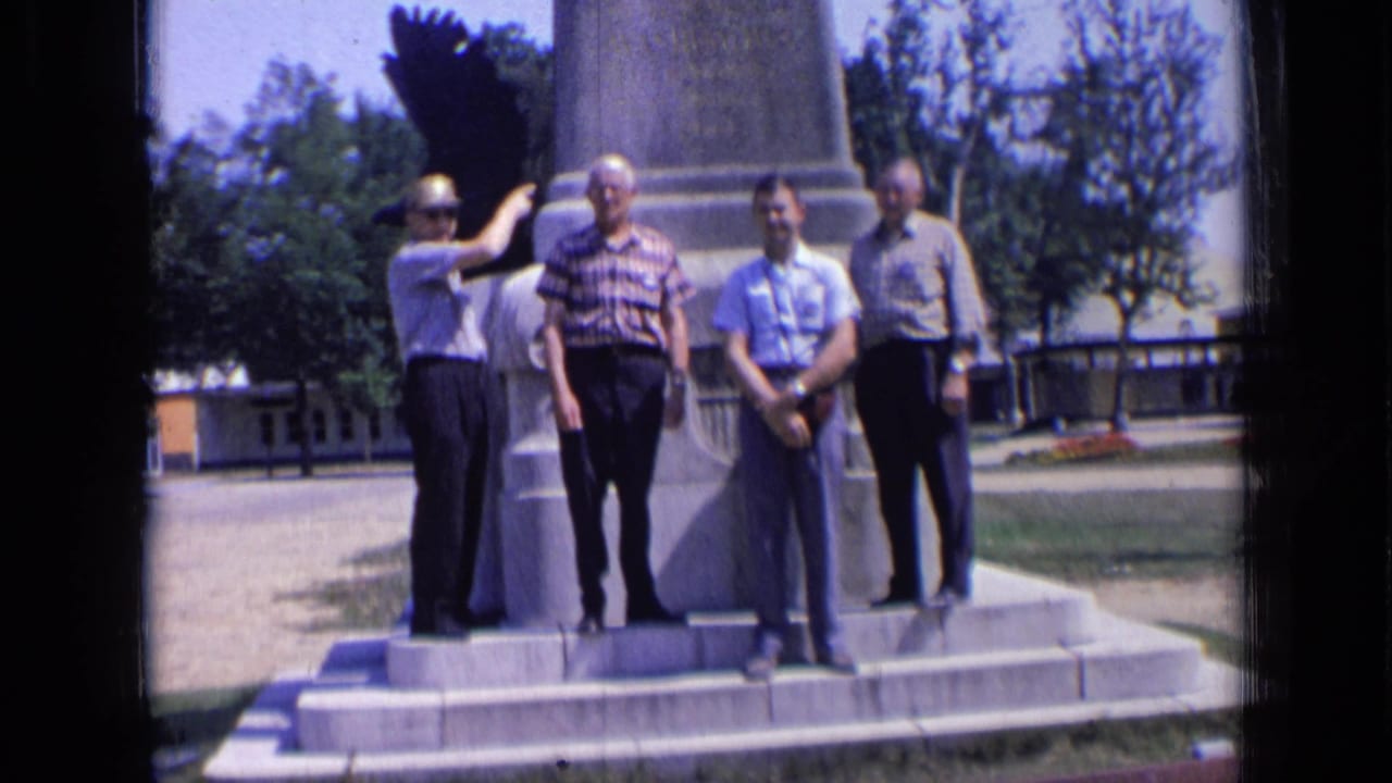 Four adult men in summer shirts and trousers pose at the base of a large monument in Russia, with one man pointing toward the structure.