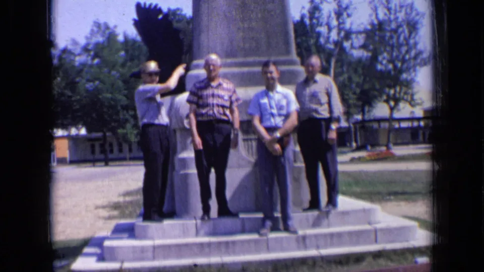 Four adult men in summer shirts and trousers pose at the base of a large monument in Russia, with one man pointing toward the structure.