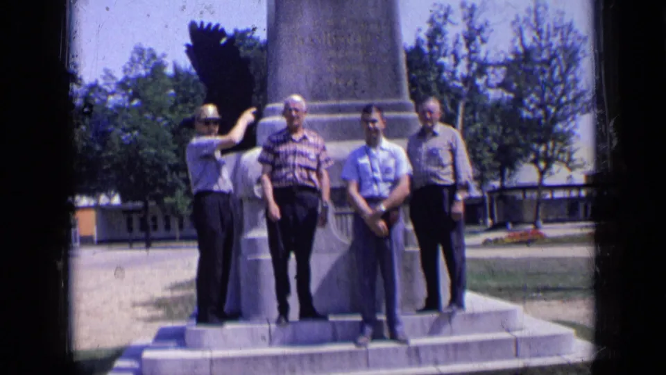 Four adult men stand on stone steps at a public monument in Russia, seen in soft 8mm color with trees and open park space behind.
