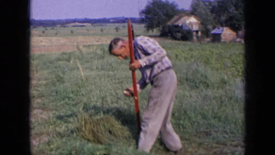 1963 Russia home-movie frame of a man working low over cut grass in an open field, with rural buildings in the background.